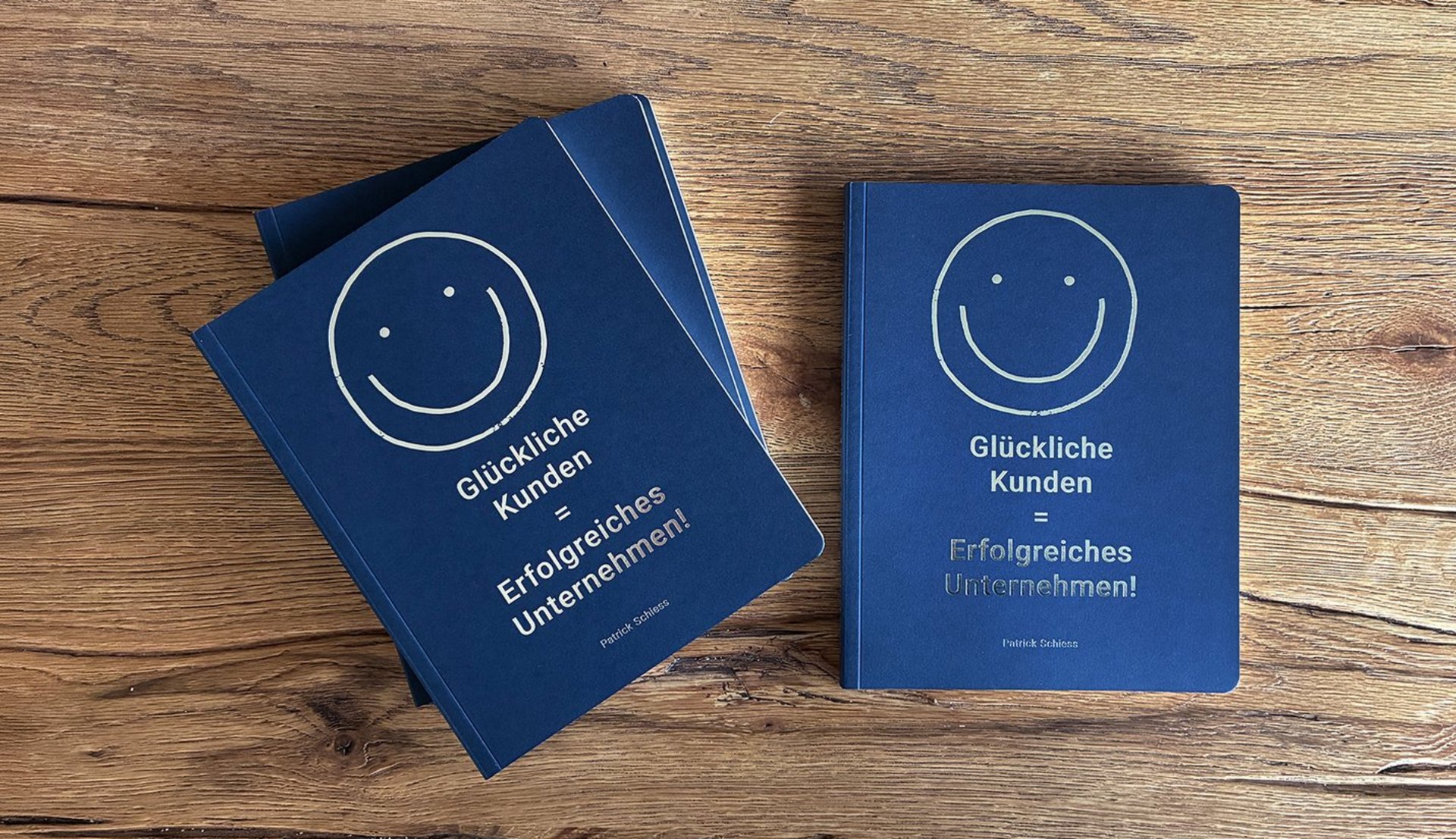 Drei dunkelblaue Bücher mit Smiley-Cover «Glückliche Kunden = Erfolgreiches Unternehmen!» liegen auf einem rustikalen Holztisch, zwei gestapelt links, eines rechts.